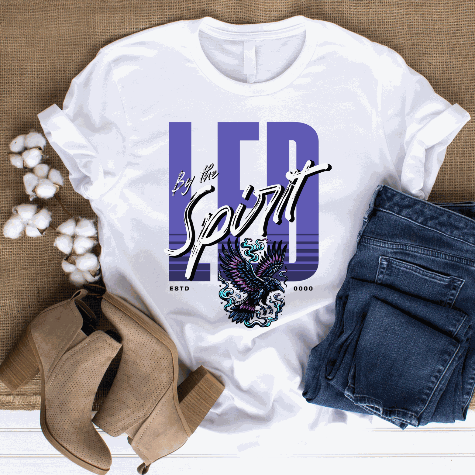 White Christian LED Unisex T-shirt with "LED by the Spirit" text and eagle graphic, paired with blue jeans and tan boots on burlap background