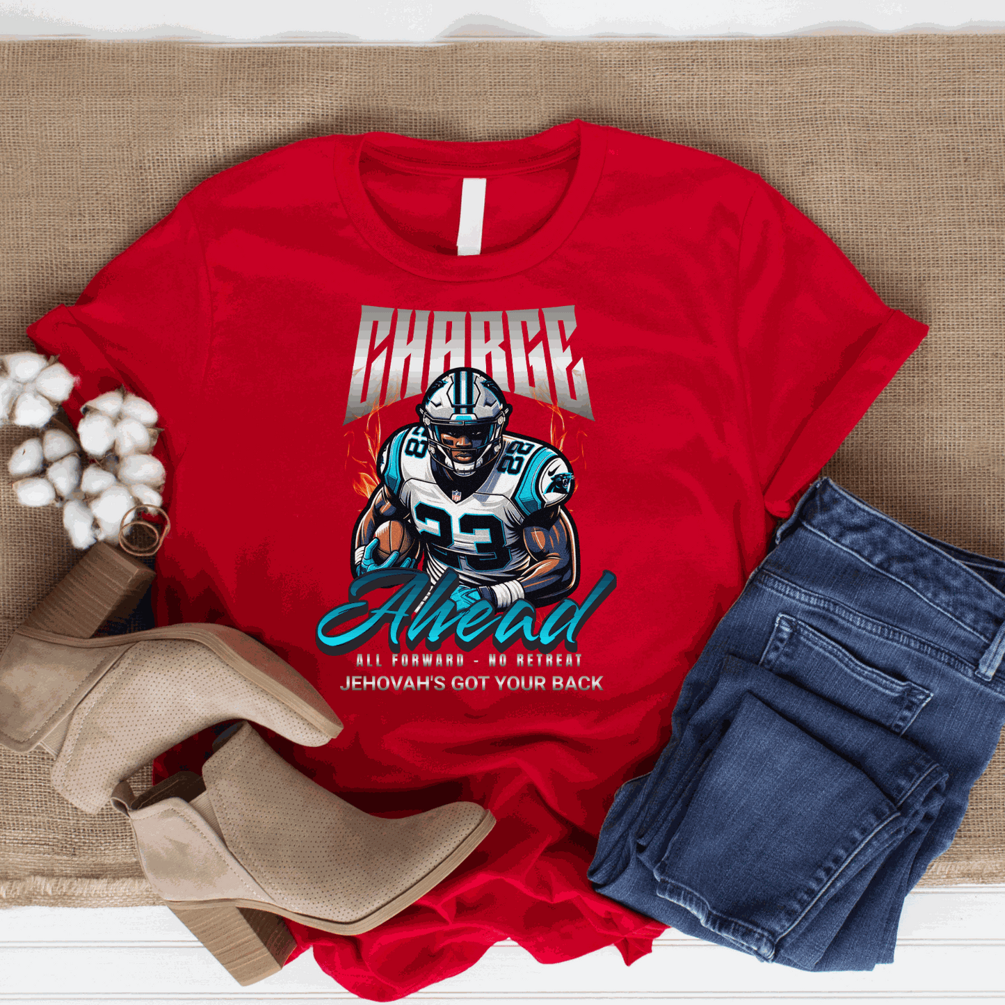 Red Charge Ahead Unisex softstyle T-shirt with football player graphic and motivational text, styled with blue jeans and beige ankle boots on burlap background.