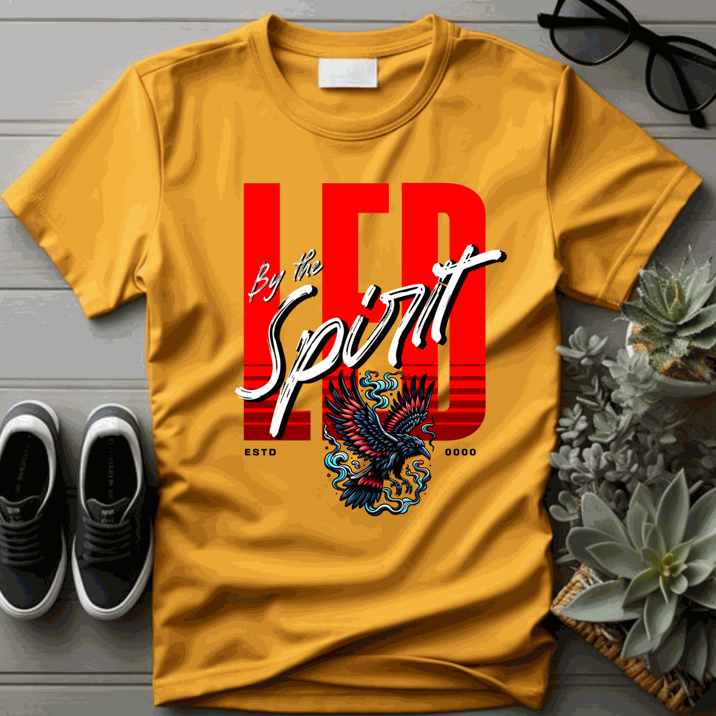 Christian LED unisex t-shirt in mustard yellow with bold red "LED" and "By the Spirit" text featuring an eagle graphic, styled with casual accessories.