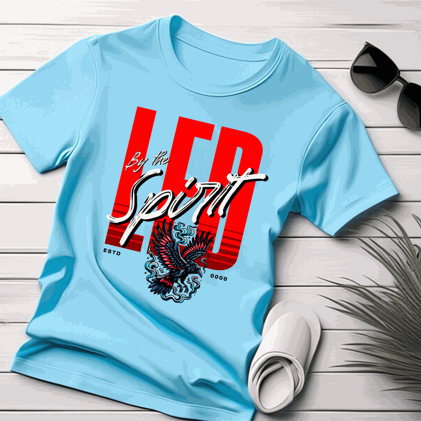 Christian LED Unisex T-Shirt in light blue with red and white "LED by the Spirit" text and eagle graphic, laid on wooden surface with sunglasses and sandals.